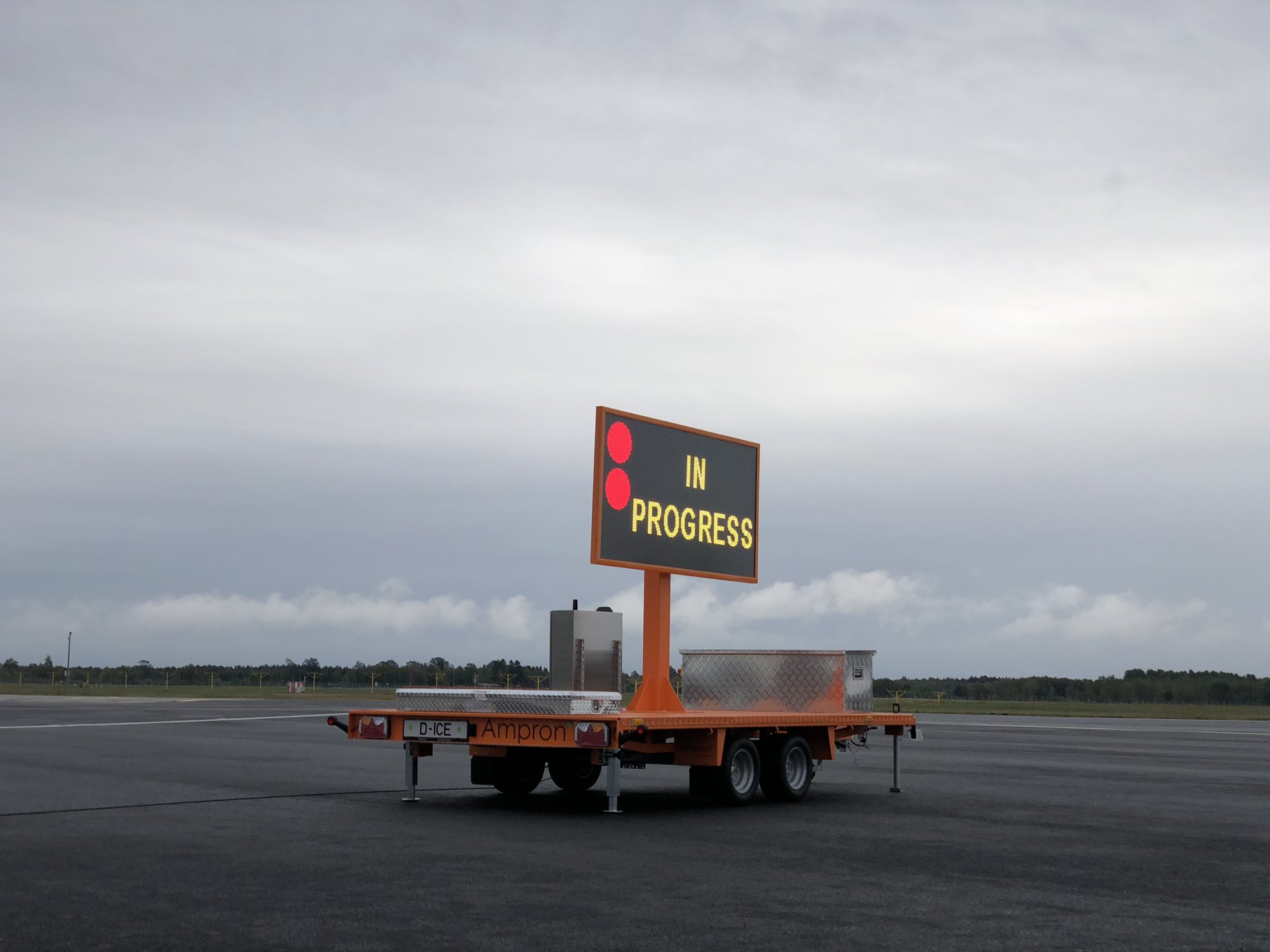 Ampron deployable LED sign on an orange trailer on the Tallinn Airport de-icing apron, showing IN PROGRESS with two red signal lights.